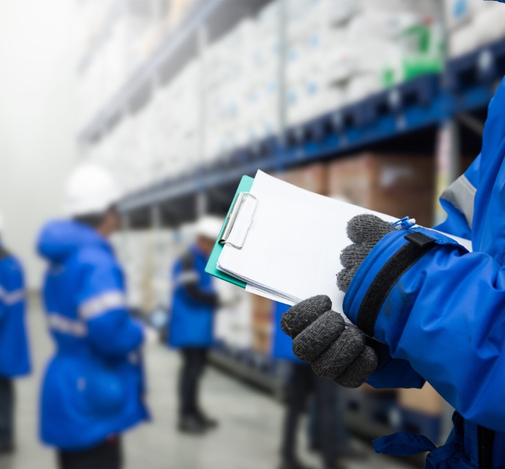A worker in a blue jacket and gloves writing on a clipboard in a warehouse.